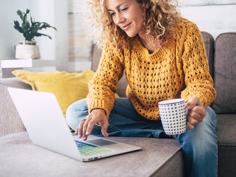 Image of woman sitting on couch browsing on her laptop with coffee in hand.