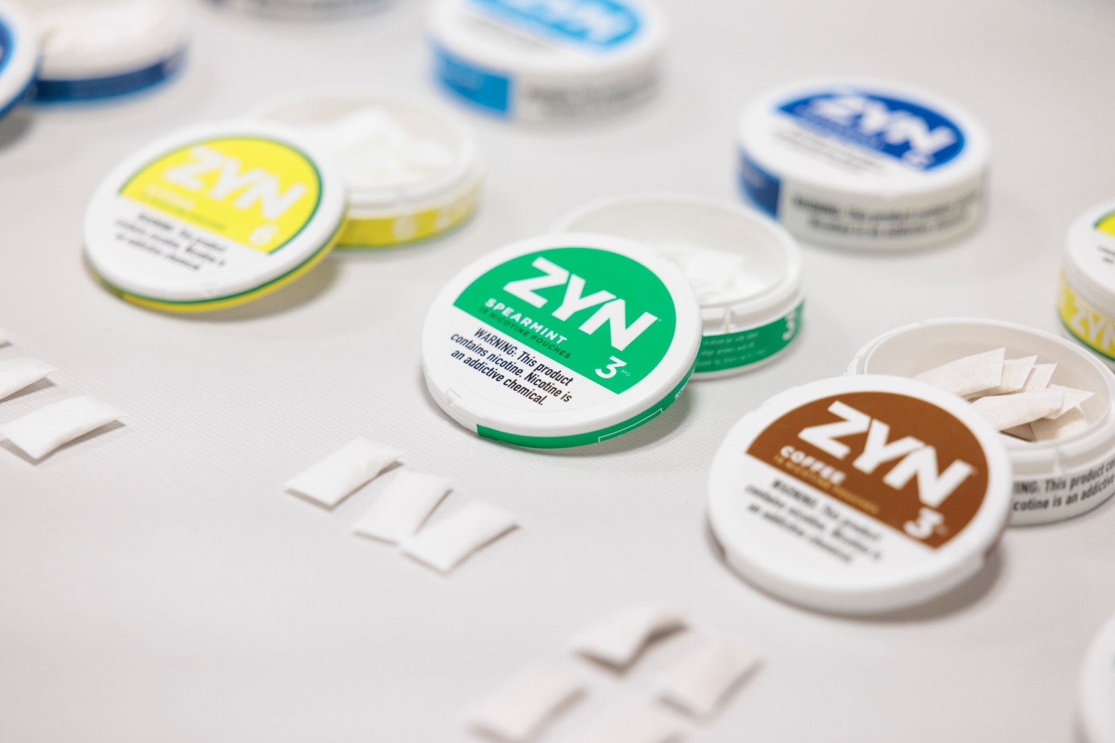 The image shows a selection of ZYN nicotine pouch products arranged on a table. The cans are prominently displayed, each featuring different flavours such as spearmint and coffee, clearly labeled with a nicotine strength of 3 mg. The background includes scattered individual pouches, emphasizing the variety and usage of the product. The overall setting suggests a display or promotional event for these tobacco-free nicotine alternatives.