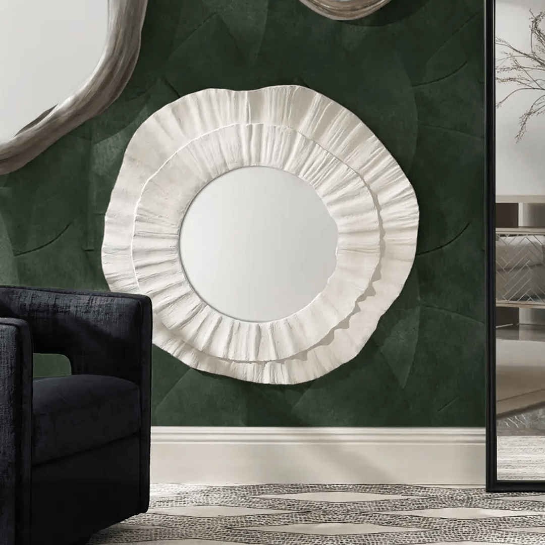 Striking interior scene showcasing a Z Gallerie mirror with a bold, sculptural white frame resembling layered petals or ruffled fabric, mounted on a deep green textured wall. The organic round mirror is paired with a modern black accent chair and framed by glimpses of adjacent decor, creating a dramatic and artistic focal point.