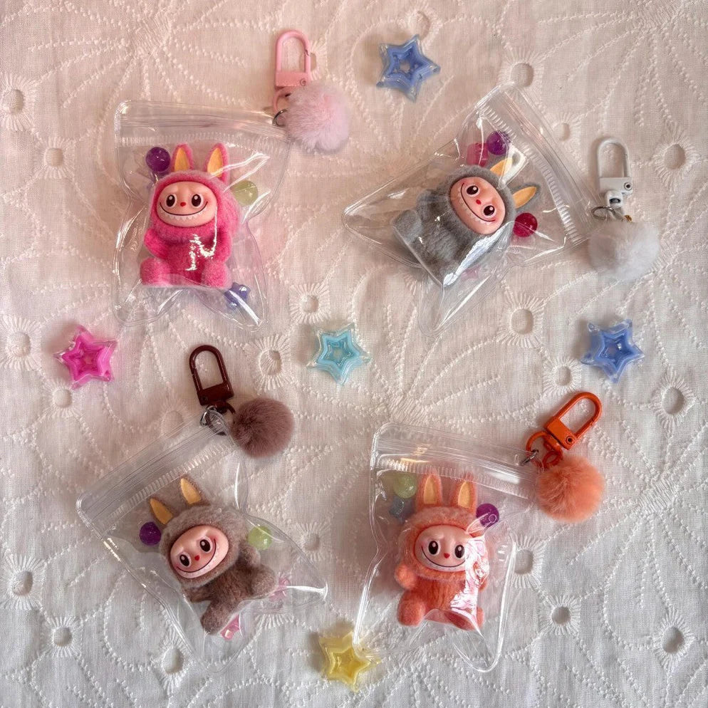 Assorted Labubu character keychains in clear zip pouches, each with a colorful clip and pom-pom charm, displayed on a textured white fabric with decorative star beads.