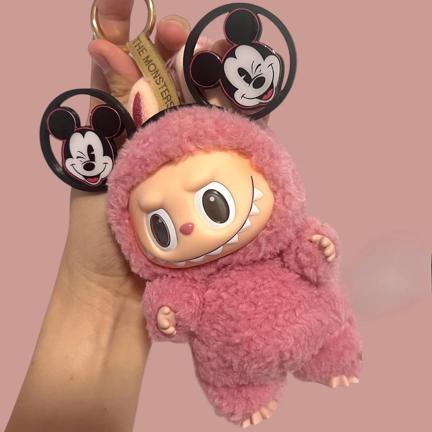 Hand holding a pink fuzzy Labubu plush keychain dressed in a textured suit, with Mickey Mouse-themed ear charms and a strap labeled 'The Monsters'.