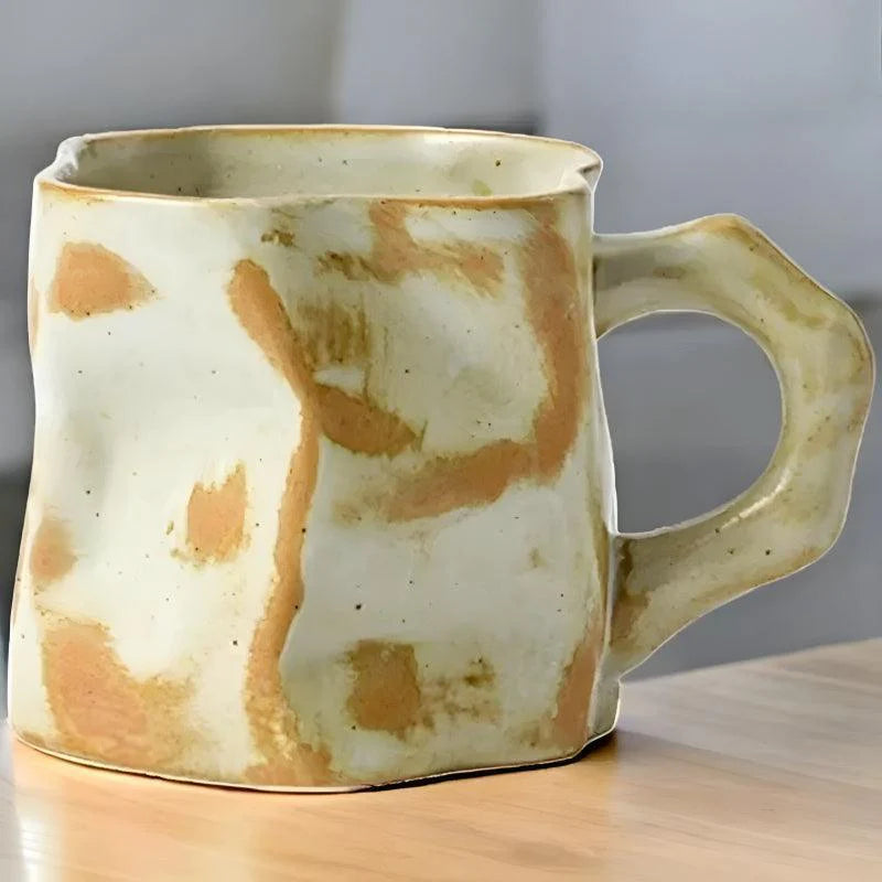 A uniquely shaped ceramic mug with an abstract orange and cream pattern, featuring a chunky angular handle, placed on a wooden surface.
