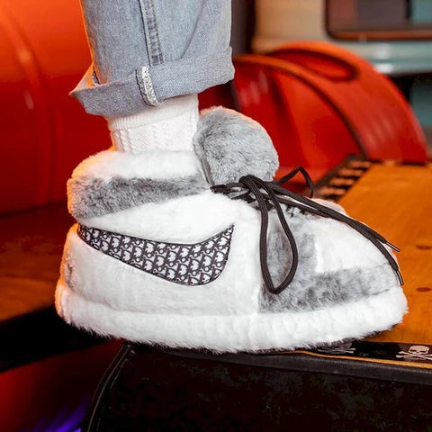 Person wearing plush gray and white sneaker-style slippers with black laces and patterned side detail.