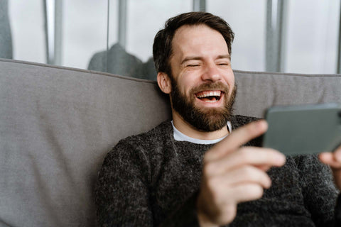handsome bearded man in black woolen sweater, sitting on grey couch laughing at a joke